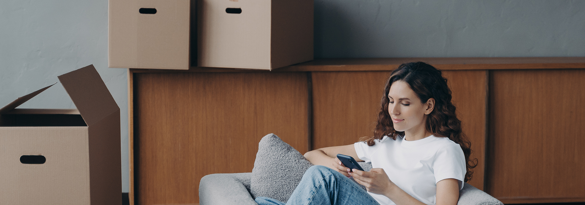 A képen egy hölgy ül egy kamapén, mobiltelefonját nézi, körülötte dobozok vannak a szobában