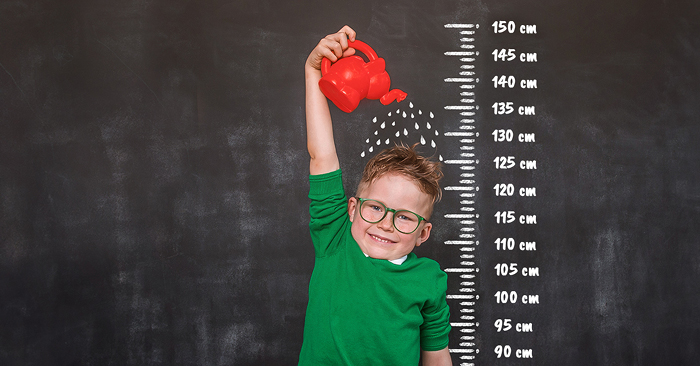 A képen egy kisfiú áll egy falra krétával rajzolt mérőszalag előtt és locsolja magát egy piros locsolókannával