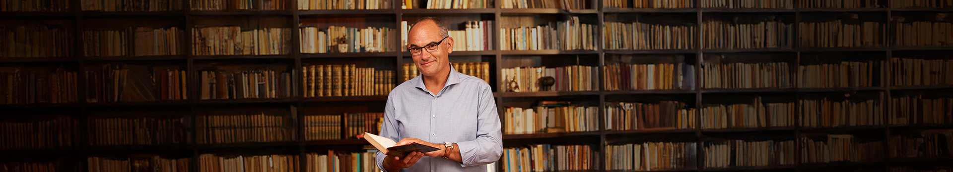 Gundel Takács Gábor könyvet lapozgat, háttérben könyvespolcok.